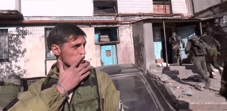 Mikhail Tolstykh looking afar while smoking with 3 army persons entering the house in the background. Mikhail wearing a brown jacket, green tactical vest, and bracelet