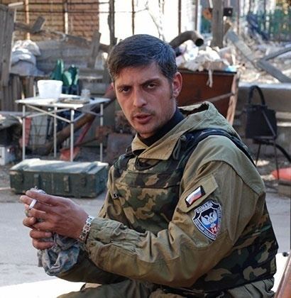 Mikhail Tolstykh holding a cigarette, with a serious face and mustache, while wearing a brown jacket, camouflage tactical vest, and bracelet