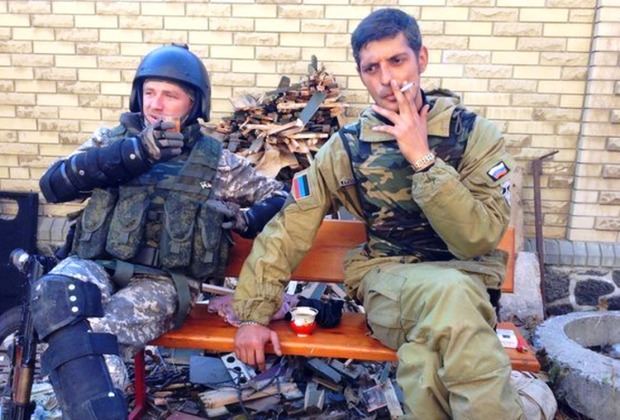 Mikhail Tolstykh is smoking and the soldier beside him is holding a glass of juice while they are sitting on the bench. Mikhail wearing a brown jacket, pants, and camouflage vest