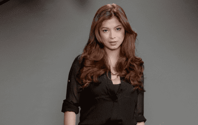 Angel Locsin with a serious face, wavy long chestnut-colored hair on a dark gray background, wearing a black long sleeve blouse.