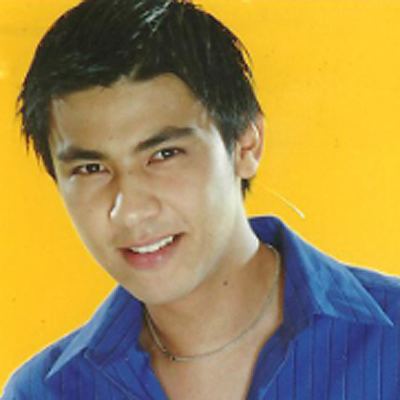 Miko Sotto smiling while looking at something, black hair with a mole on his neck on a yellow background, wearing a necklace and a blue striped collared shirt.