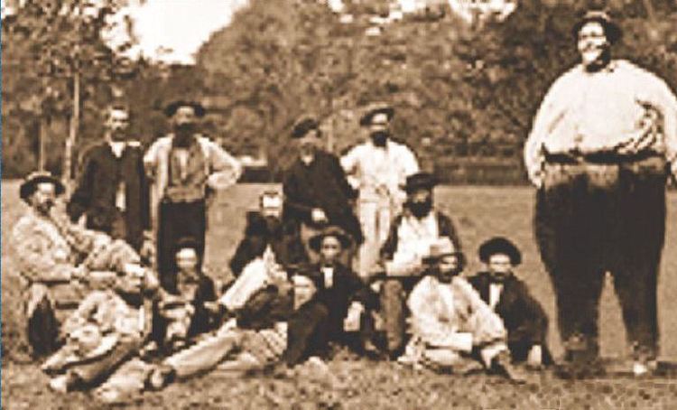 Mills Darden (right) standing with a group of people and some of them are sitting on the grass while others are standing, with trees in the background. Mills wearing a hat, shoes, a long sleeve, and pants with a belt