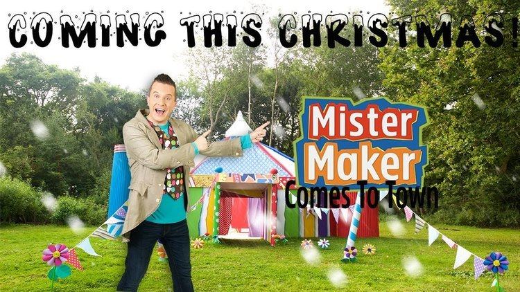 Poster of Mister Maker Comes to Town featuring Phil Gallagher with a big smile and pointing his fingers at the logo, wearing a khaki coat over a polka dot printed vest and blue-green shirt.