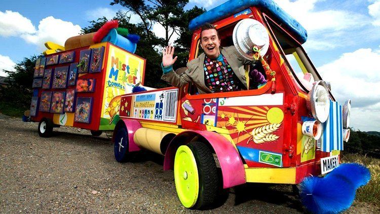 Phil Gallagher with a big smile while waving his hand and riding in the Mister Maker mobile, wearing a khaki coat over a polka dot printed shirt.