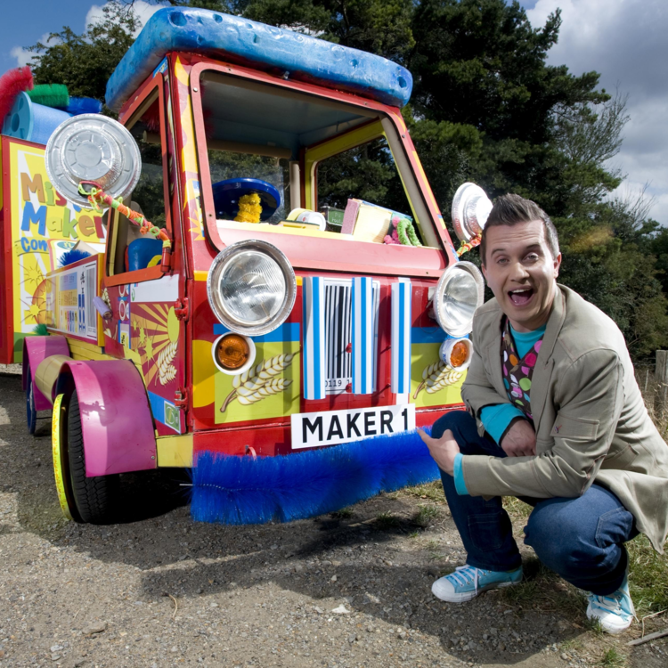 Phil Gallagher with a big smile and pointing his fingers at Mister Maker's mobile, wearing a khaki coat over a polka dot printed vest and blue-green shirt, jeans, and blue shoes.