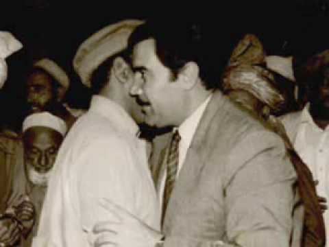 A man whispering to Mohammad Najibullah while being surrounded by people, and he is wearing a coat, long sleeves, and necktie
