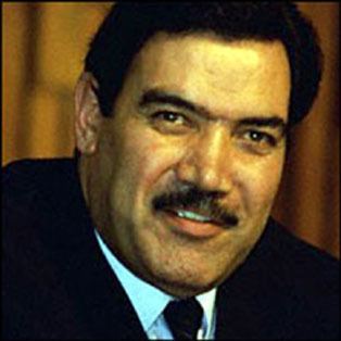 Mohammad Najibullah smiling while wearing a black coat, white long sleeves, and black necktie