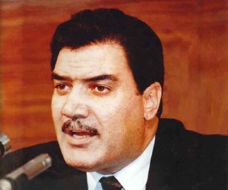 Mohammad Najibullah speaking using a microphone while wearing a black coat, white long sleeves, necktie
