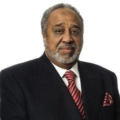 Mohammed Hussein Al Amoudi's tight-lipped smile while wearing a black coat, white long sleeves, and red necktie