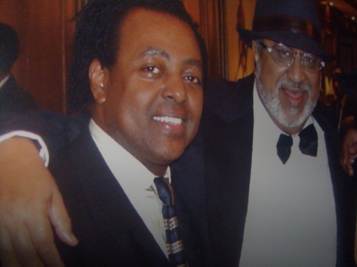 Mohammed Hussein Al Amoudi smiling with the man beside him while wearing a black hat, eyeglasses, black coat, and black bow tie