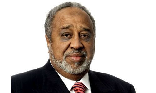 Mohammed Hussein Al Amoudi's tight-lipped smile while wearing a black coat, white long sleeves, and red necktie