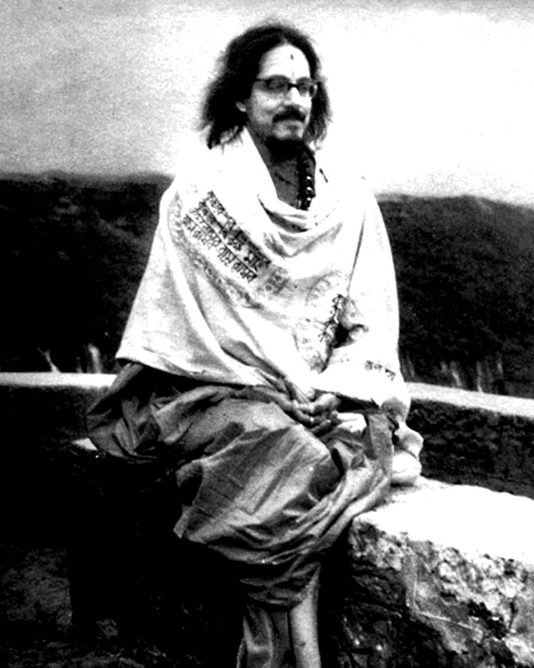Mohanananda Brahmachari sitting on the bench while he has long hair and a mustache and wearing a shawl, eyeglasses, and dress