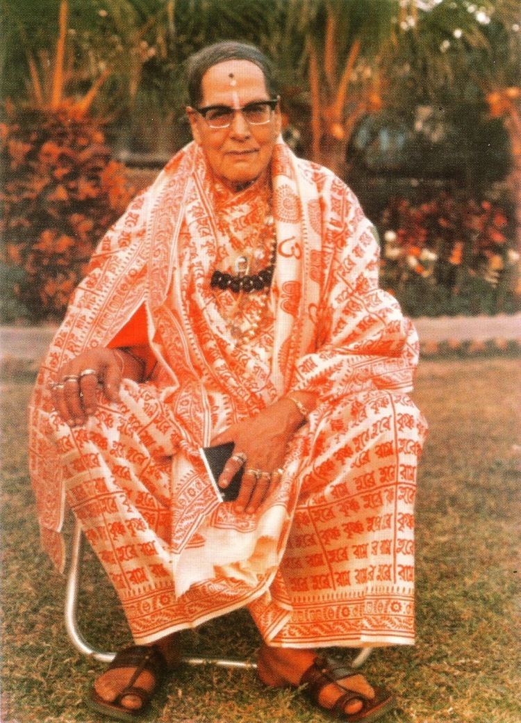 Mohanananda Brahmachari smiling while sitting on the chair and wearing a white and orange robe, black necklace, rings, and eyeglasses