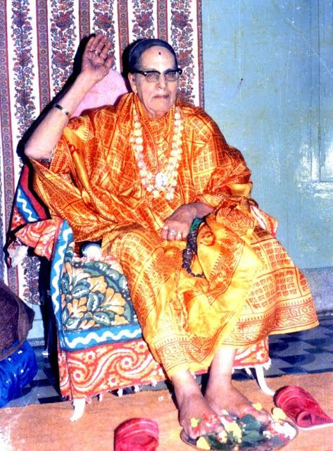 Mohanananda Brahmachari smiling while sitting on the chair and wearing a yellow and orange robe, necklace, rings, bracelet, and eyeglasses