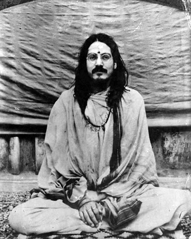 Mohanananda Brahmachari sitting on the floor while he has long hair and a mustache and wearing a shawl, eyeglasses, and dress