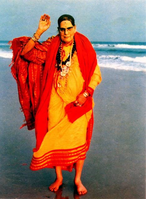 Mohanananda Brahmachari waving his hand while at the seashore and wearing a yellow and orange dress, shawl, eyeglasses, and some jewelry