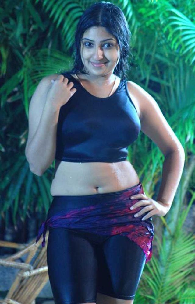 Monica smiling while hand on her hips and wearing a black sleeveless crop top, black short leggings, and a cloth wrap around her waist