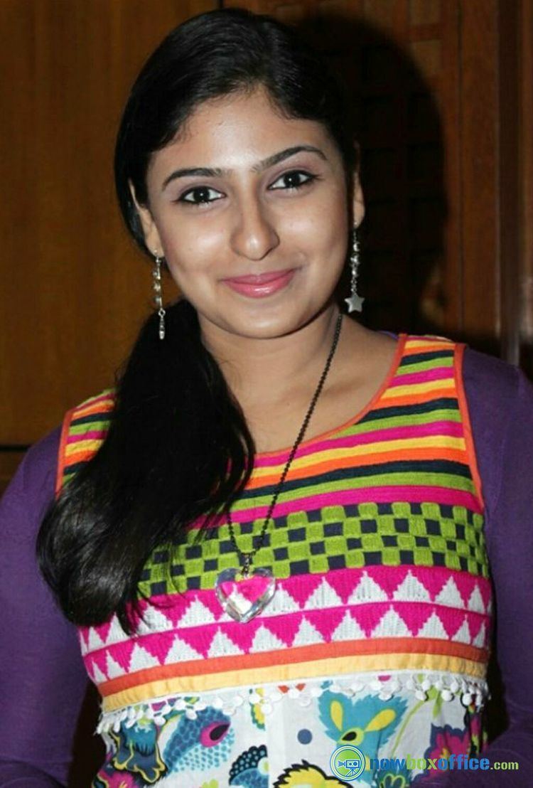 Monica with a tight-lipped smile while wearing a colorful long sleeve blouse with a different patterns, heart necklace, and earrings
