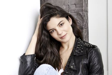 Monica Barbaro is smiling and sitting while touching her hair with her right hand,  and wearing a black leather jacket and denim pants
