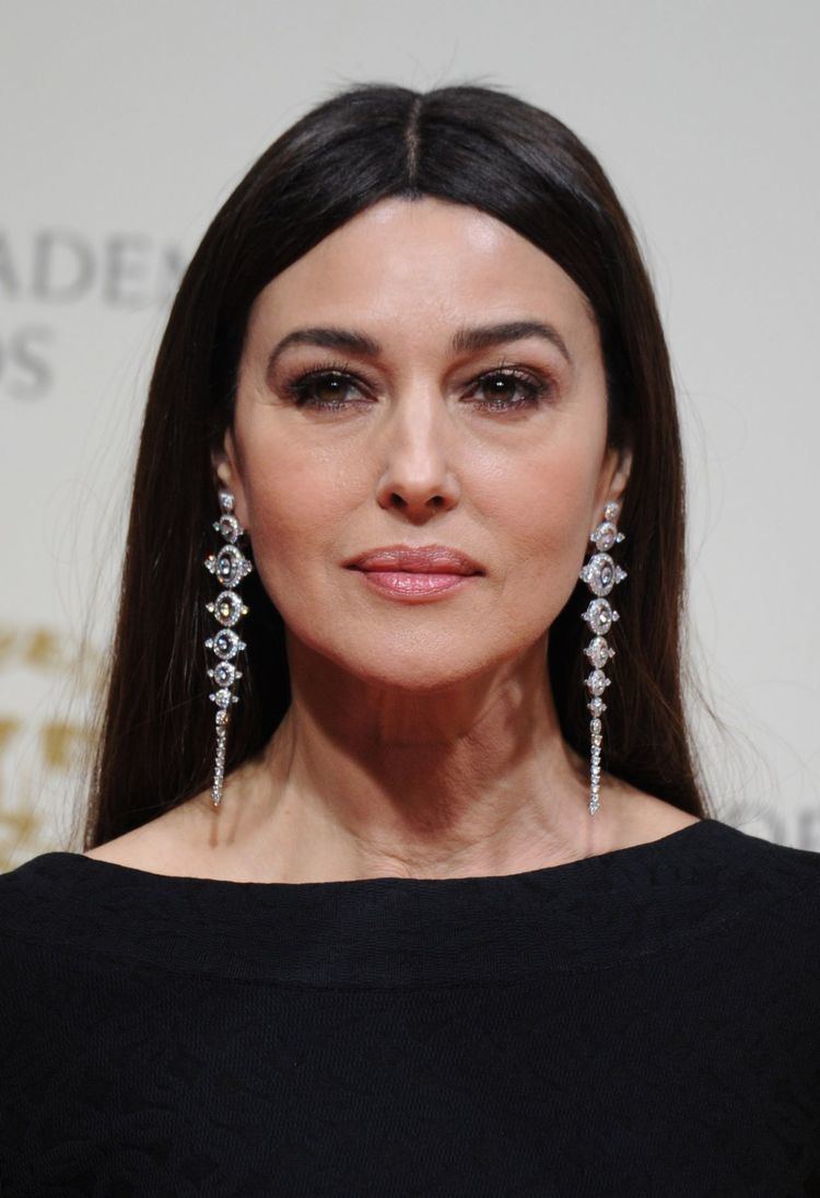 Monica Bellucci looking afar and wrinkles on her neck are visible while wearing shoulder-duster earrings and a black blouse