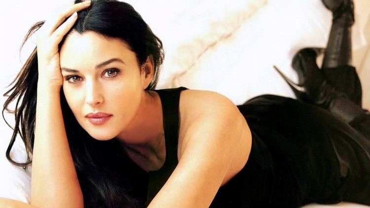 Monica Bellucci lying on the bed while hand on her head and wearing a black sleeveless top, black pants, and black boots