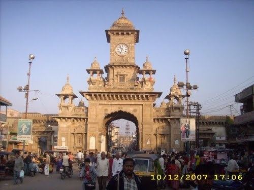 The Nehru gate clock tower at Morbi Gujarat India Asia has large tall concrete walls with unique patterns, at the top is a large clock with a round and pointy roof. Has a crowd of people in front of the gate with large tall speakers and street lights, along with the transportation vehicles on the road at the right bottom is the date 10/12/2007.