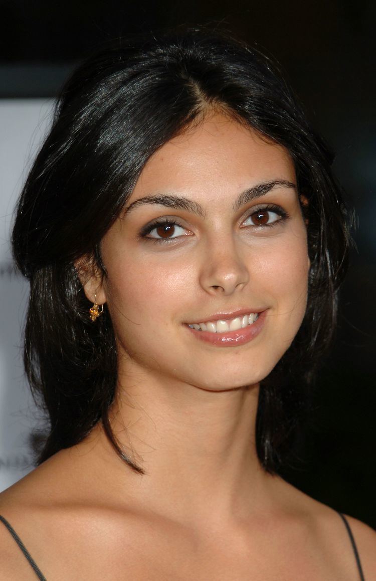 Morena Baccarin smiling with black shoulder-length hair while wearing earrings and a spaghetti top with black strap