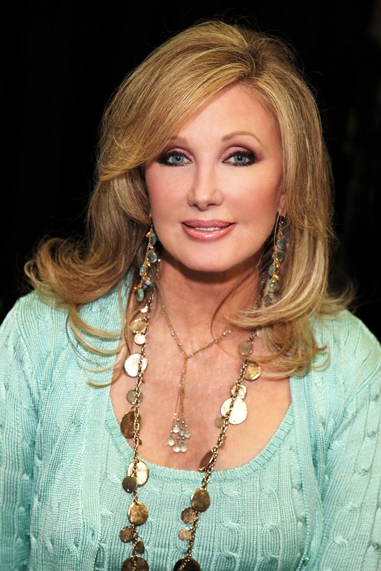Morgan Fairchild smiling while wearing a green knitted blazer, green knitted blouse, gold layered necklace, and earrings