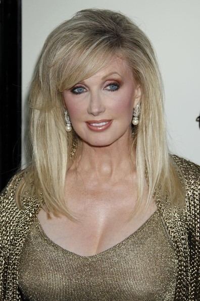 Morgan Fairchild smiling while wearing a gold and black blazer, gold inner blouse, and earrings