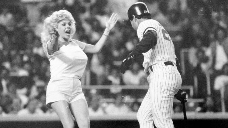 Morganna wearing a white shirt and shorts with a baseball player wearing his baseball uniform inside a baseball court.