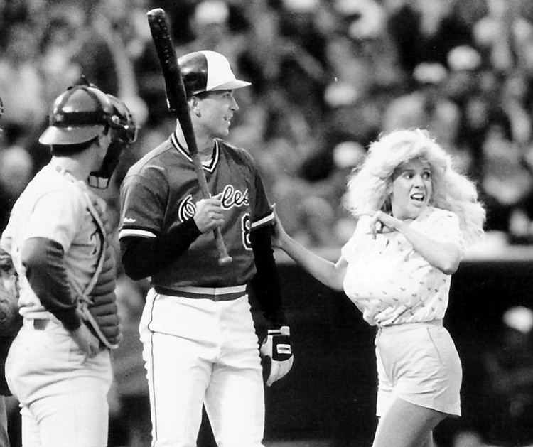 Morganna wearing a shirt and shorts with two baseball players wearing their baseball uniforms inside a baseball court.