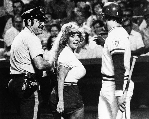 A policeman in his uniform, wearing a cap and eyeglasses escorting Morganna wearing a white shirt and shorts with a baseball player holding a baseball bat inside a baseball court.