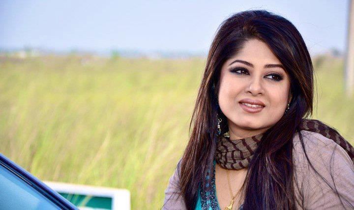 Moushumi smiling while looking afar and wearing a brown striped scarf, beige blazer, blue blouse, and necklace