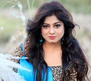 Moushumi with a tight-lipped smile and black curly hair while wearing blue and leopard print blouse and blue earrings