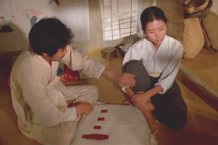 In the movie scene of Mulberry (film) 1986, in a room with wooden walls with painted cloth from left, a man is sitting on the ground legs crossed, while holding Lee’s arm with his left hand, has black hair wearing a white korean robe and pants, at the right, Lee Mi-sook is serious, sitting on the ground right knee up holding with his right arm while holding it with her left hand, has black hair wearing a white long sleeve top and a dark green pants.