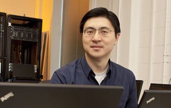 Mung Chiang is smiling, has a short mustache, and black straight hair, with two laptops in front and a system unit behind on his right side, wearing eyeglasses, and a white t-shirt under black polo long sleeves.
