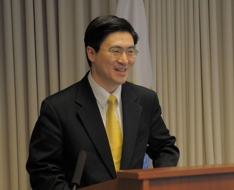 Mung Chiang is smiling with his teeth, has a short mustache, and black straight hair, he is standing in front of a microphone, wearing eyeglasses, and white long sleeves with a yellow necktie under a black coat.