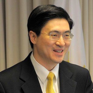 Mung Chiang is smiling with his teeth, has a short mustache, black straight hair, wearing eyeglasses, white long sleeves with a yellow necktie paired with a black suit.