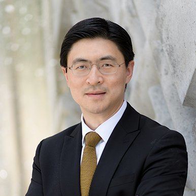 Mung Chiang is smiling, has a short mustache, black straight hair, wearing eyeglasses and white long sleeves with a brown necktie under a black coat