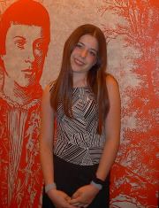 Jennifer Ann Crecente smiling and standing in front of a red portrait of a man while wearing a black and white striped sleeveless blouse and bracelet