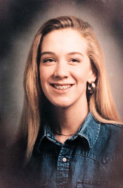 Leslie Mahaffy smiling with golden-brown hair and braces on her teeth while wearing hoop earrings, a necklace, and a blue denim blouse with a collar.