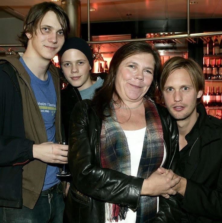 My Skarsgård smiling together with her sons, Sam, Valter, and Bill. My Skarsgård with long hair, wearing a white shirt and a multi-colored scarf.