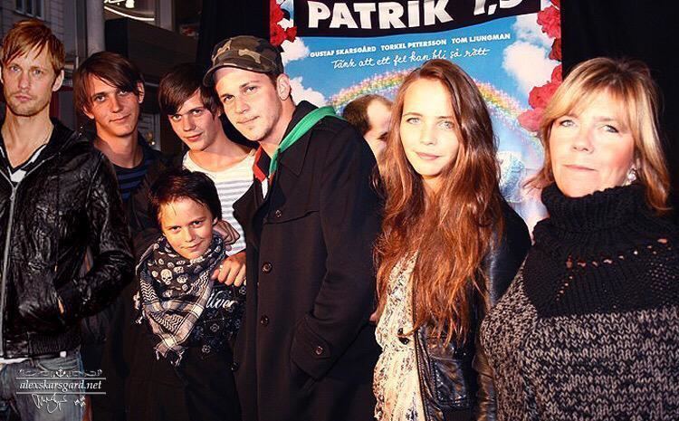 My Skarsgård smiling together with her six children Alexander, Gustaf, Valter, Eija, Sam, and Bill. My Skarsgård with blonde hair, wearing a black knitted scarf around her neck and a black top.