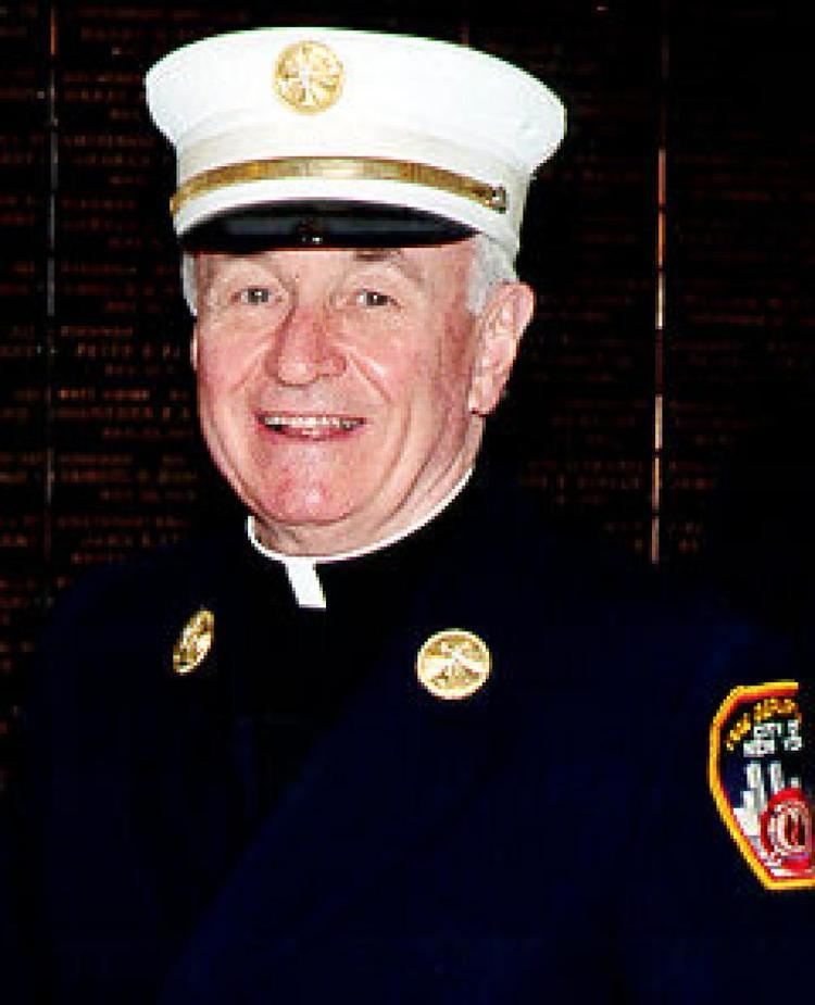 Mychal Judge smiling while wearing a peaked cap, a blue coat with badges, and black long sleeves