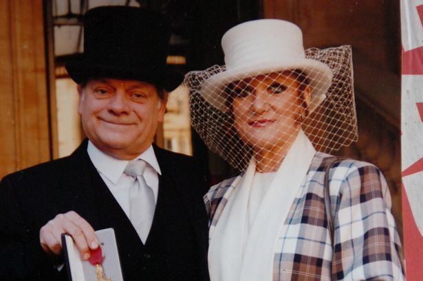 Myfanwy Talog wearing hat with veil, checkered coat and white inner top and David Jason wearing black hat, black coat, white long sleeves and gray neck tie as he shows his OBE