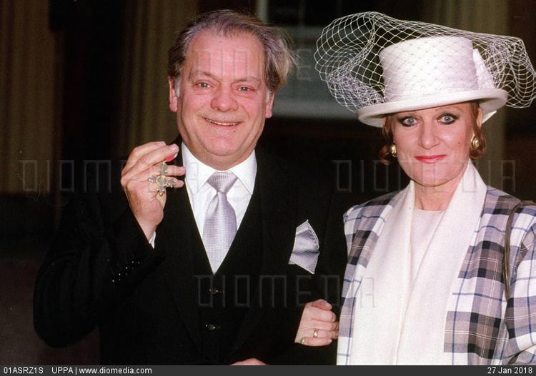 Myfanwy Talog wearing hat with veil, checkered coat and white inner top and David Jason wearing black hat, black coat, white long sleeves and gray neck tie as he shows his OBE