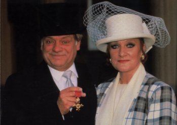 Myfanwy Talog wearing hat with veil, checkered coat and white inner top and David Jason wearing black hat, black coat, white long sleeves and gray neck tie as he shows his OBE