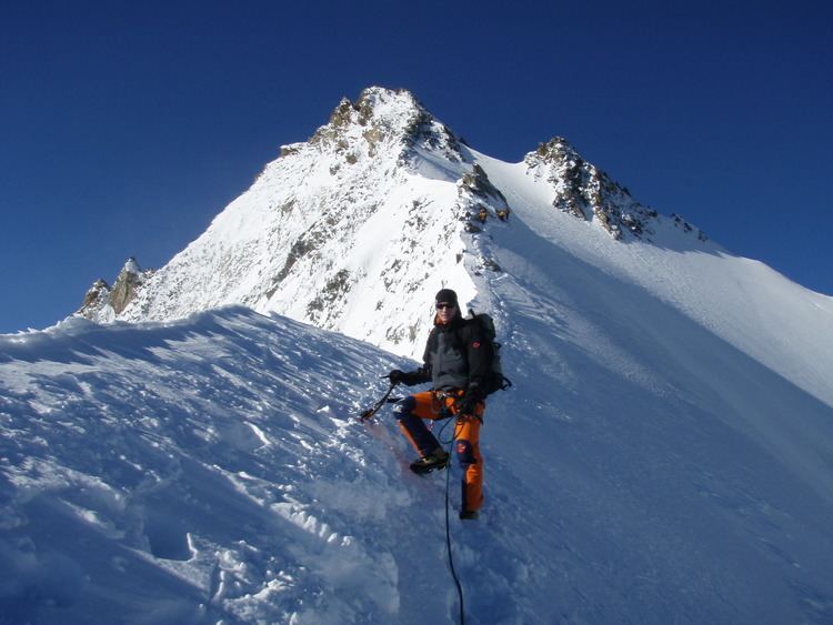 A man posing at Mount Nadelhorn while holding a harness and carrying a black backpack and wearing a black beanie, black jacket, sunglasses, and black and orange pants