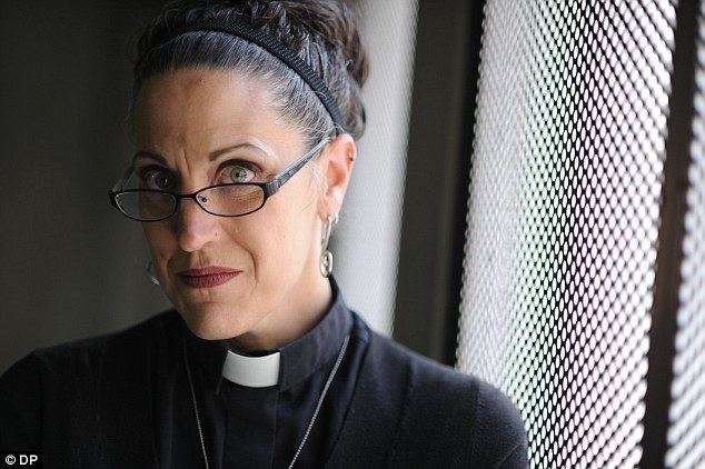 Nadia Bolz-Weber posing inside a confession room wearing a black pastors outfit, a headband and a rosary.
