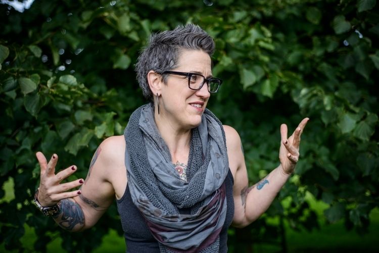 Nadia Bolz-Weber explaining something while having a standing hairstyle and wearing a sleeveless shirt and a thick scarf.